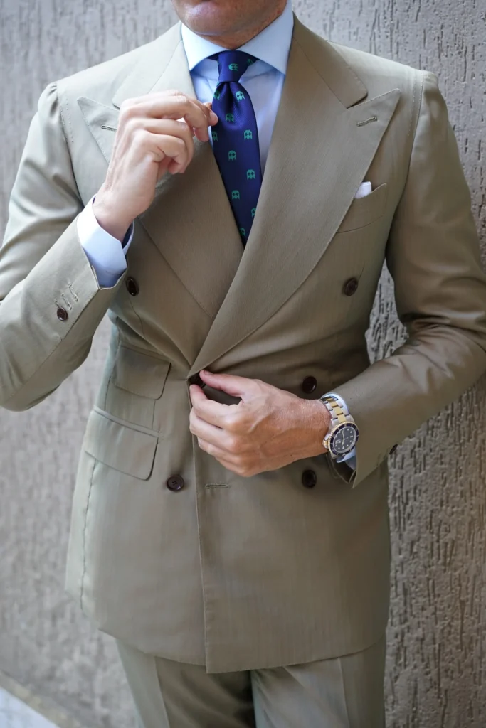 Un homme élégant ajuste sa cravate bleue à motifs, vêtu d’un costume croisé beige parfaitement taillé, incarnant un style chic et raffiné.