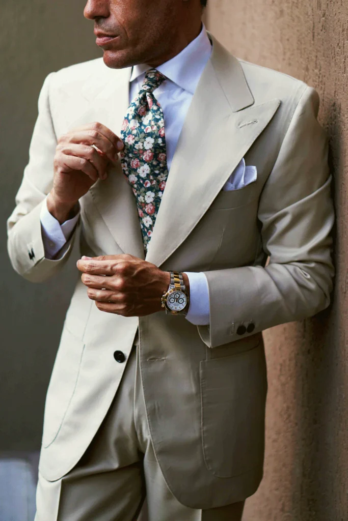 Élégance intemporelle : un costume clair parfaitement taillé, rehaussé d’une cravate florale et d’une montre classique. Un style qui murmure le raffinement.