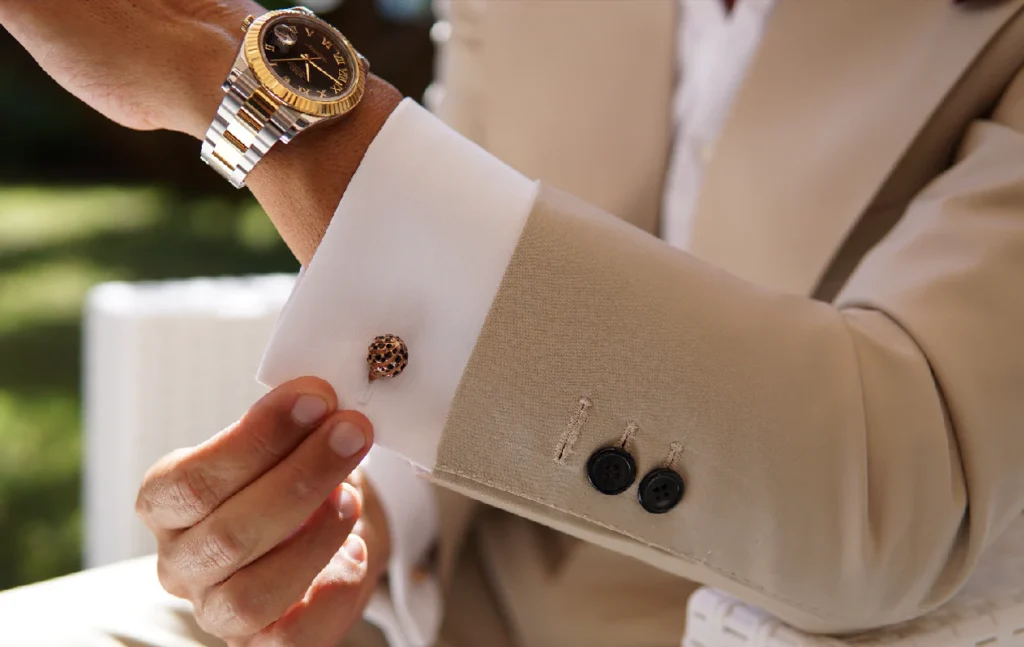 Gros plan raffiné d’un poignet habillé d’un costume beige clair, associé à une chemise blanche à poignets mousquetaires. Un bouton de manchette élégant, aux finitions métalliques travaillées, apporte une touche de distinction subtile. La montre-bracelet bicolore en acier et or renforce l’allure luxueuse de l’ensemble, tandis que les boutons sombres de la manche et la coupe nette du costume soulignent une élégance masculine soignée et contemporaine.
