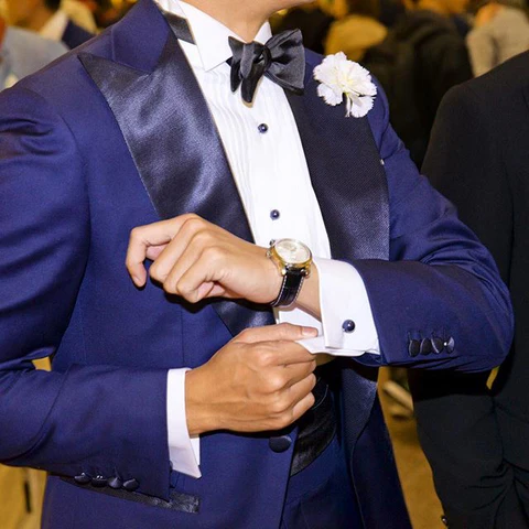 Cette photographie met en avant une variante audacieuse du smoking classique avec un ensemble bleu nuit profond, où le cummerbund en soie noire crée un contraste texturé sous la veste à revers en pointe satinés. L'accessoire assure une transition parfaite entre le pantalon et la chemise de cérémonie à plastron plissé, soulignant l'importance des détails comme le nœud papillon coordonné et la boutonnière florale pour un look de gala moderne. L'ajustement du poignet révèle également des boutons de manchette élégants, prouvant que le cummerbund reste la pièce maîtresse pour structurer une silhouette masculine haut de gamme.