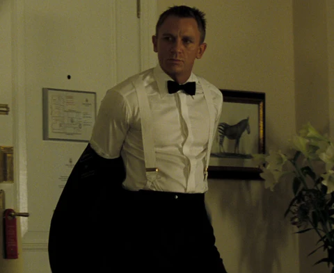 Dans cette scène iconique, Daniel Craig dans le rôle de James Bond illustre les dessous de l'élégance formelle en portant des bretelles blanches sur une chemise de soirée impeccable, juste avant d'enfiler sa veste. Bien que le cummerbund ne soit pas encore visible ici, la taille haute du pantalon noir et le nœud papillon déjà noué préparent le terrain pour cet accessoire traditionnel qui viendra parfaire sa tenue d'agent secret. Cette image souligne que la rigueur du style "Black Tie" repose sur une superposition méticuleuse d'éléments, où chaque pièce, des bretelles à la ceinture de smoking, contribue à une allure de distinction intemporelle.