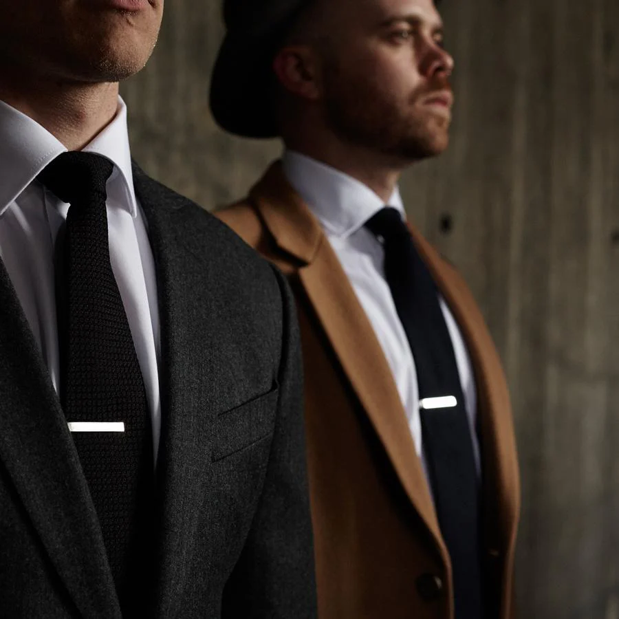 Deux hommes portant des tenues formelles avec des cravates sombres, chacun utilisant une pince à cravate argentée pour un look soigné.