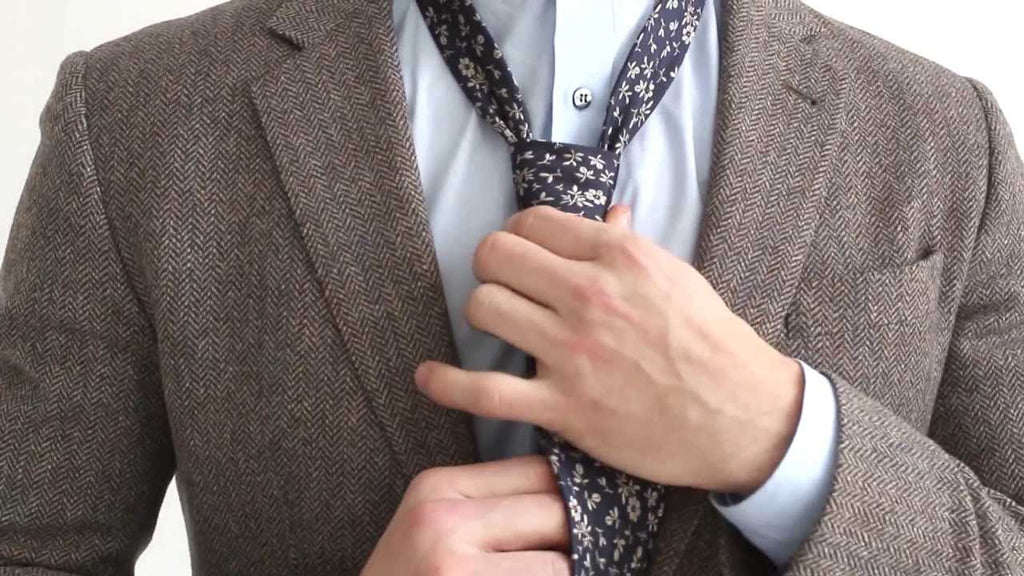 Un homme en train d'ajuster soigneusement le nœud de sa cravate à motifs floraux discrets sur une chemise bleue et une veste en tweed.