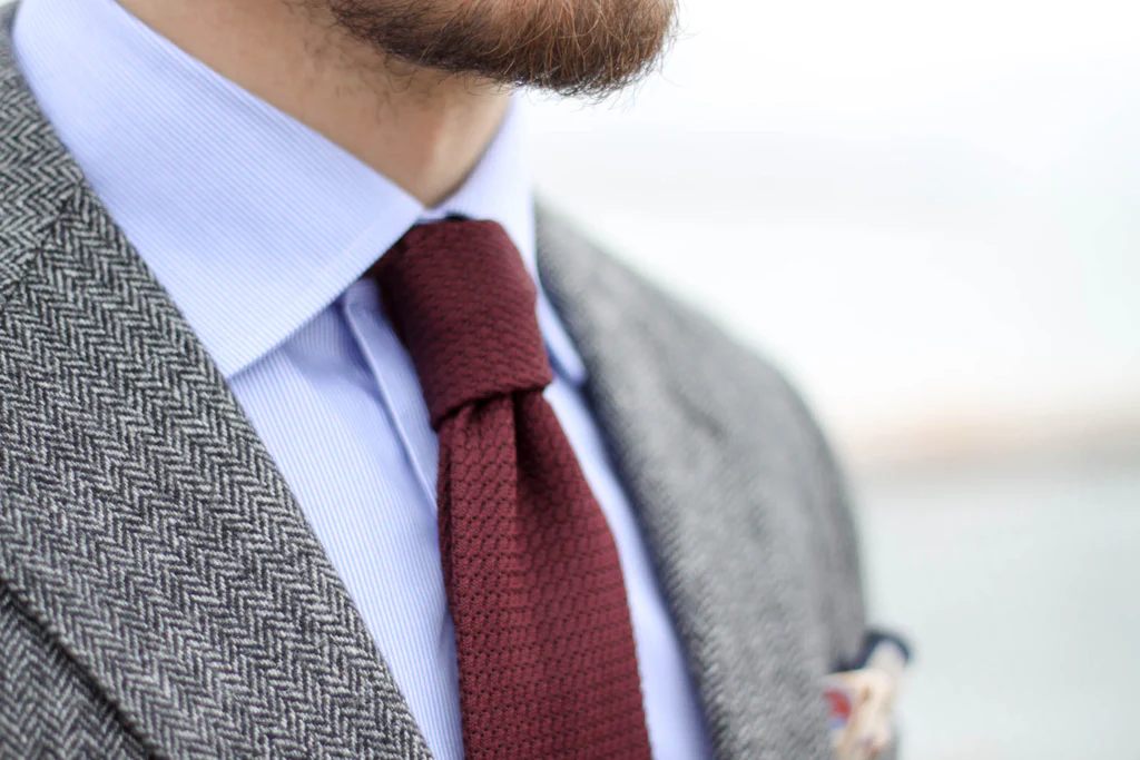Un plan rapproché mettant en valeur la texture d'une cravate en tricot bordeaux, portée avec une veste à chevrons grise.