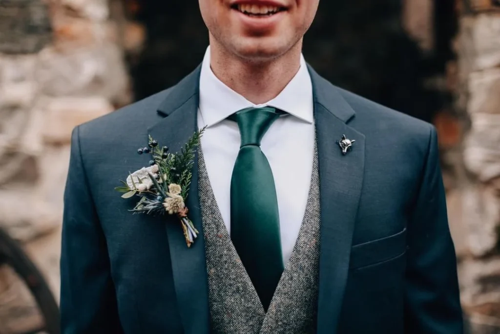 Costume bleu profond porté avec un gilet texturé et une cravate vert foncé, accompagné d’une boutonnière végétale inspirée de la nature. Une tenue de marié moderne qui allie authenticité, finesse et charme intemporel.