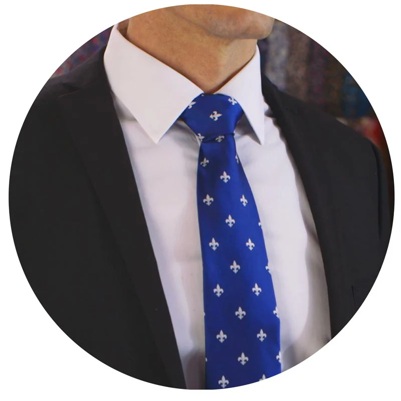 Un homme portant un costume sombre avec une chemise blanche et une cravate bleue à motif fleur de lys blancs. La cravate est nouée de manière soignée et symétrique, donnant un style élégant et formel.